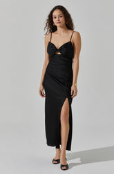 A woman is wearing a long, black satin dress with spaghetti straps and a knot detail at the bust. The dress features a cut-out below the bust and a high slit on the left side, revealing her leg. She has accessorized with gold hoop earrings, a gold bracelet, and black open-toe heels.